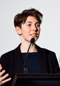 Image of Cat Nadel speaking into a microphone at a lectern with a white background