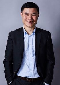 Image of Tim Lo Surdo smiling at the camera in a black suit jacket with his hands in his pocket, with a grey background